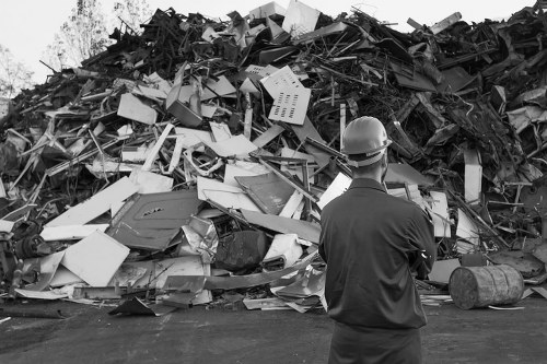 A man wearing a safety helmet and dark work clothes stands on a paved outdoor area, observing a large heap of mixed waste materials including broken furniture, discarded appliances, metal scrap, and various debris. The waste pile is densely packed, with items piled high against a backdrop of trees and partly visible structures. The materials show a variety of textures and finishes, such as smooth plastic, rough metal, and chipped wood, in shades of white, black, and metallic. The scene appears to be at a waste or recycling yard, illustrating a typical rubbish removal site, possibly in the vicinity of Luton, with the man representing a waste management professional or site supervisor. House Clearance Luton offers services to remove and clear such waste, ensuring proper disposal and clearance of clutter from residential or commercial properties. The natural lighting highlights the complexity of the waste, emphasizing the importance of professional rubbish removal services for environmental and safety reasons.