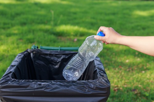 A person disposing of a clear plastic water bottle into a black open-top rubbish bin on a grassy outdoor area, with green lawns and blurred natural background. The hand, visible from the wrist, is holding the bottle by its neck, and the bottle appears to be partially crushed. The waste bin is situated on the ground, likely in a garden or residential backyard, with no other objects immediately visible nearby. The scene reflects typical rubbish disposal activity relevant to household or garden waste collection in Luton, as handled by House Clearance Luton, with the focus on responsible waste management and rubbish removal services.