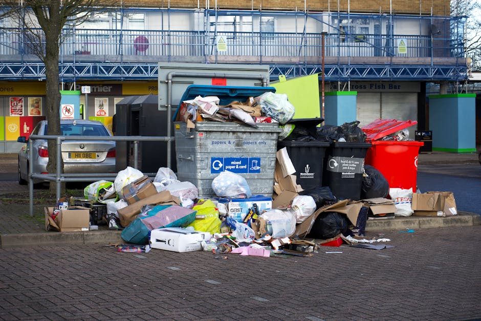 A large overflowing public rubbish bin lined with a blue lid and marked for mixed paper and cardboard, situated on a paved urban street with piles of discarded waste surrounding it. The waste includes
