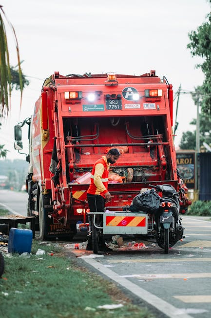 A large overflowing public rubbish bin lined with a blue lid and marked for mixed paper and cardboard, situated on a paved urban street with piles of discarded waste surrounding it. The waste includes