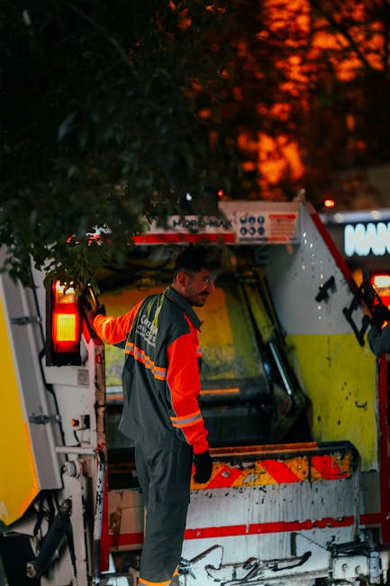 A street scene at dusk showing a waste collection vehicle with its rear compartment open, revealing a large, heavy-duty metal container used for rubbish disposal. The vehicle is painted primarily whit