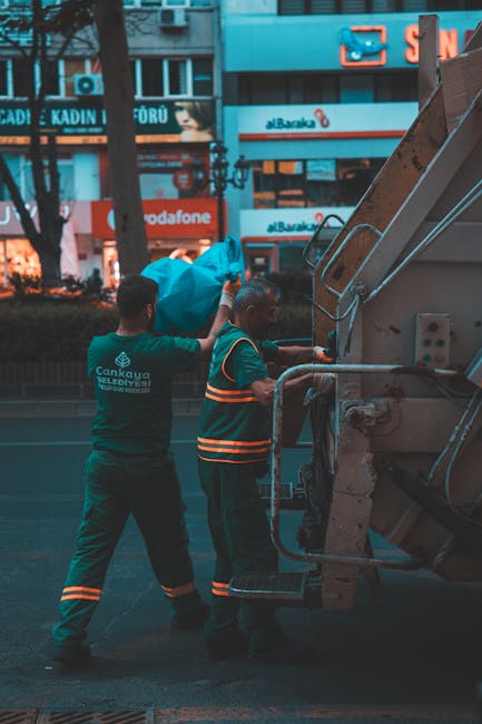 A street scene at dusk showing a waste collection vehicle with its rear compartment open, revealing a large, heavy-duty metal container used for rubbish disposal. The vehicle is painted primarily whit