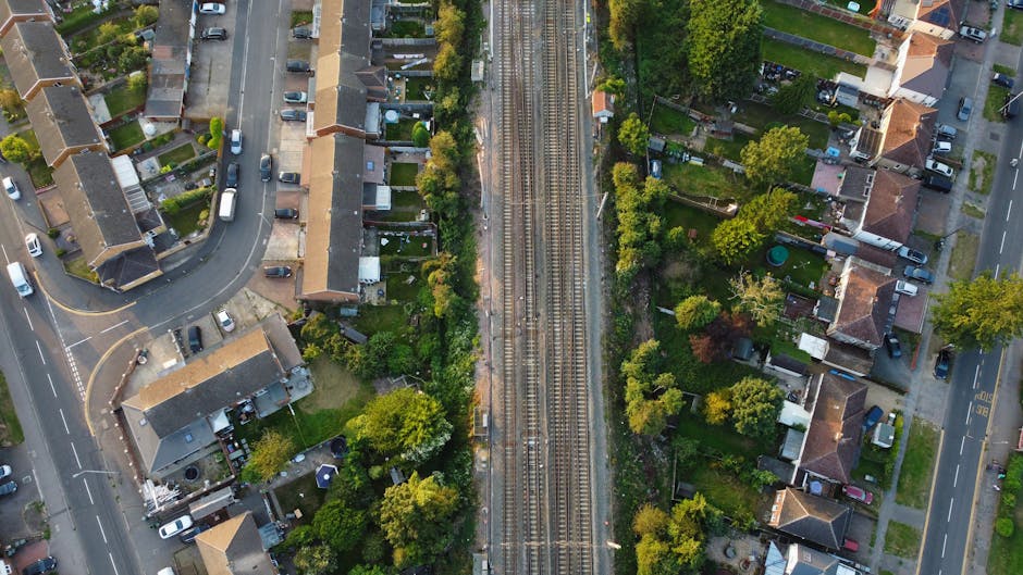 An aerial view of a suburban residential area featuring a set of railway tracks running vertically through the center of the image. On either side of the tracks are rows of detached and semi-detached